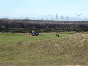 A golf course at Newbiggin Golf Club with manicured green fairways and sandy dunes in the foreground. A tractor is maintaining the course while a golfer pulls a trolley nearby. In the background, wind turbines and power lines stretch across the horizon, contrasting the natural landscape with industrial elements. 