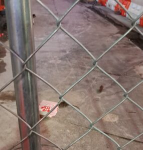 A chain-link fence in the foreground encloses a dimly lit site with a concrete floor. Several rats are visible scurrying across the ground, one near a discarded popcorn container. Orange and white safety barriers line the edges of the site, and faint tire tracks mark the dusty pavement. The scene is illuminated by artificial lighting, casting a muted glow over the area.