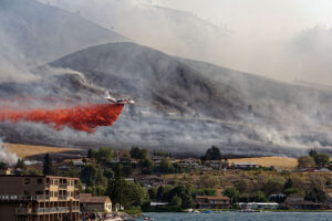 A large air plane plays drops a vivid red retarding red cloud on a smoked and burning hillside in Chelan, Washington. The forest fire, part of the fire of the Chelan complex, crawls dangerously near the residential houses located in the base of the hills. The thick smoke moves through the landscape, partially obscuring the burned terrain. The scene captures the intense effort to protect the community from advanced flames.