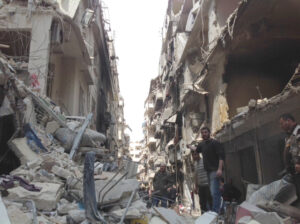 A war-damaged street in the Christian quarter of Aleppo, Syria, showing collapsed buildings with shattered facades and debris filling the roadway. Rubble, broken furniture, and twisted metal are strewn across the scene. Survivors navigate the destruction, some standing on the wreckage, while others move cautiously through the ruins. Sunlight filters through the open space where buildings once stood, casting long shadows over the devastation.