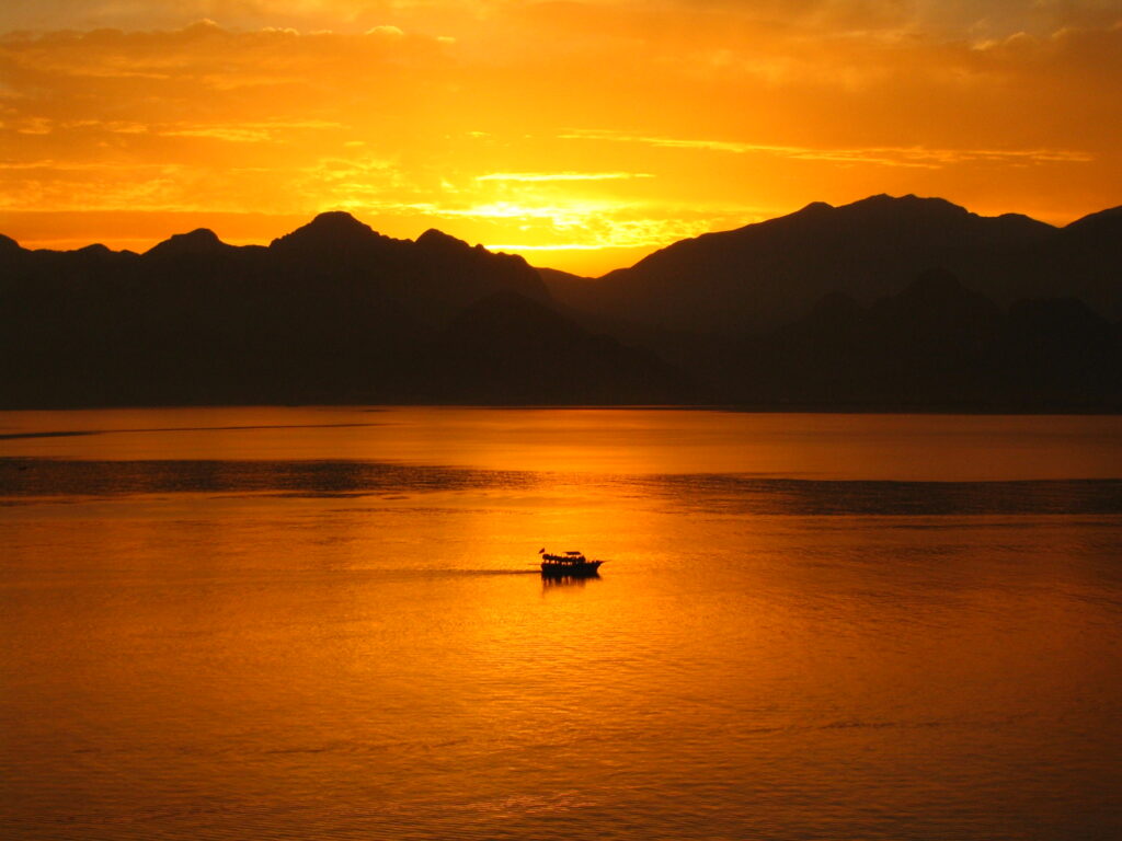 A serene sunset on a quiet body of water, with the brightness of golden orange of the sun illuminating the surface. The silhouettes form an irregular horizon in the distance, and a small boat moves peacefully through water. The scene evokes a feeling of tranquility and natural beauty.