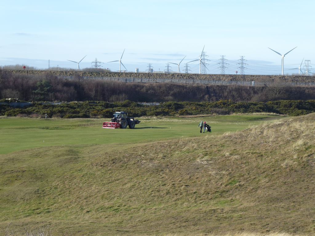 A golf course in Newbiggin Golf Club with well -maintained green streets and sandy dunes in the foreground. A tractor keeps the course while a golfer pulls a tram near. In the background, wind turbines and electric lines extend through the horizon, contrasting the natural landscape with industrial elements.