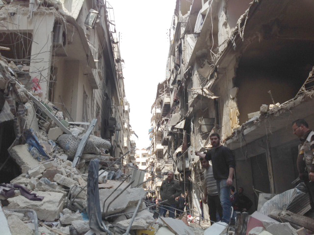 A street damaged by the war in the Christian neighborhood of Aleppo, Syria, showing buildings collapsed with shattered facades and debris that fill the road. The rubble, broken furniture and twisted metal are scattered by the scene. Survivors sail for destruction, some unemployed in the remains, while others move cautiously through the ruins. The sunlight is filtered through the open space where the buildings stopped, throwing long shadows on the devastation.